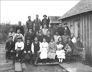 Four rows of Asian men and women sit and stand next to a one-story wooden building. Two people in front hold an infant and small child in their laps. 